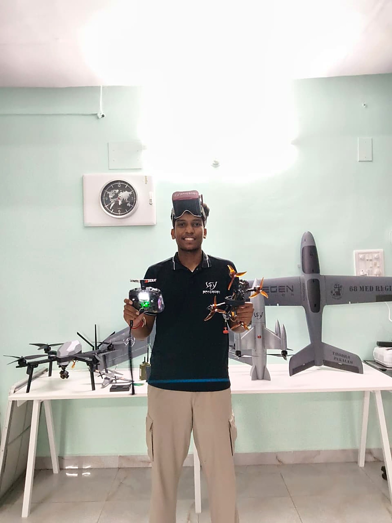 Krish Advaithan in the Sentinel Dynamics workshop, holding FPV hardware alongside VTOL and multirotor platforms.